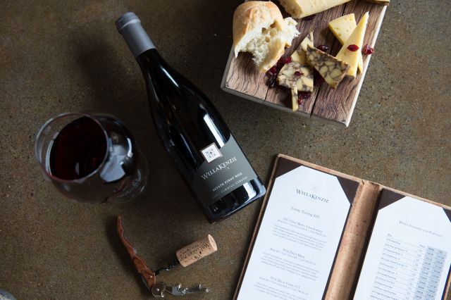 A bottle of wine laying down with a tasting menu, cheese plate. and a glass of wine laid around it.